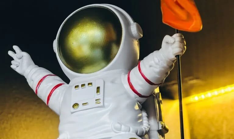 indian astronaut Shubhanshu Shukla floating inside the International Space Station wearing a white Axiom-4 mission suit with the Indian flag patch on his arm. He is smiling at the camera, with Earth visible through the Cupola window in the background. A small toy swan (zero-gravity indicator) floats beside him.
