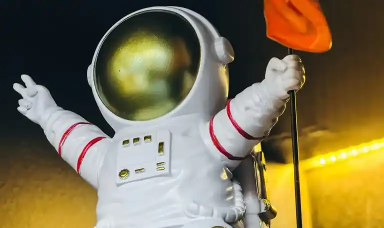 indian astronaut Shubhanshu Shukla floating inside the International Space Station wearing a white Axiom-4 mission suit with the Indian flag patch on his arm. He is smiling at the camera, with Earth visible through the Cupola window in the background. A small toy swan (zero-gravity indicator) floats beside him.
